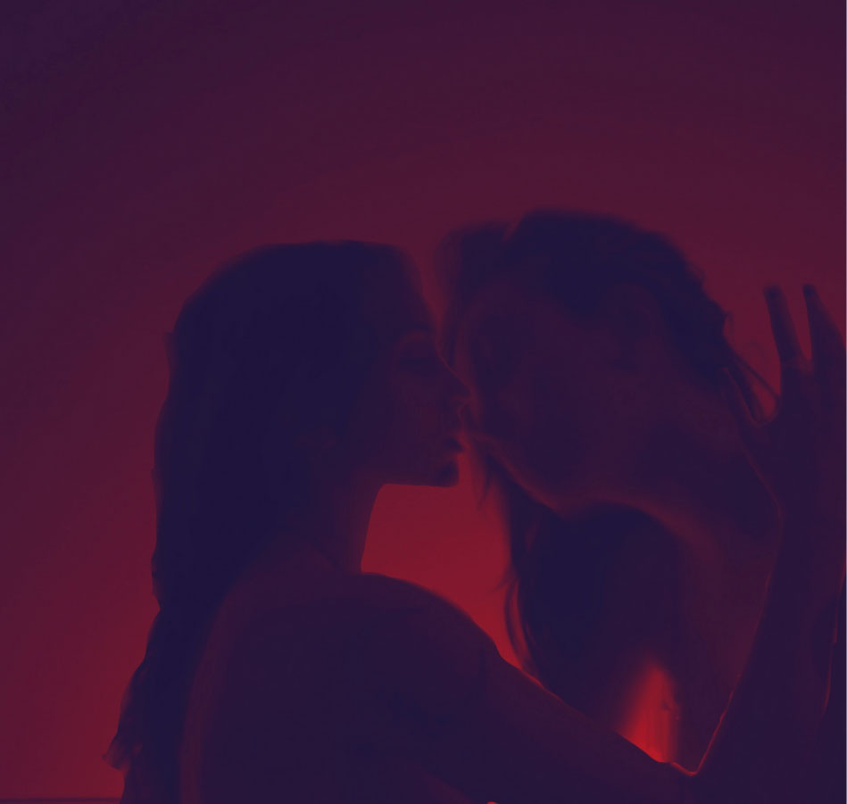 Image displays the heads of two subjects, facing each other, with their hands reaching for the other’s face. A dark orange light encircles them, while the border of this image remains darkened for dramatic effect. The subjects are in motion to simulate a kiss.