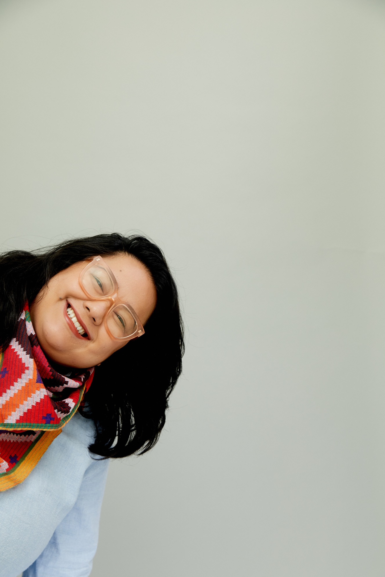 Alia is a Filipina woman standing in front of a grey background. She has black, shoulder-length hair, and is wearing glasses with light pink, transparent frames. She is wearing a light blue shirt and a scarf made from a textile that is red, white, orange, blue, green, and yellow. She is smiling and leaning in from the left side, as if she’s peeking her head in to say hello.