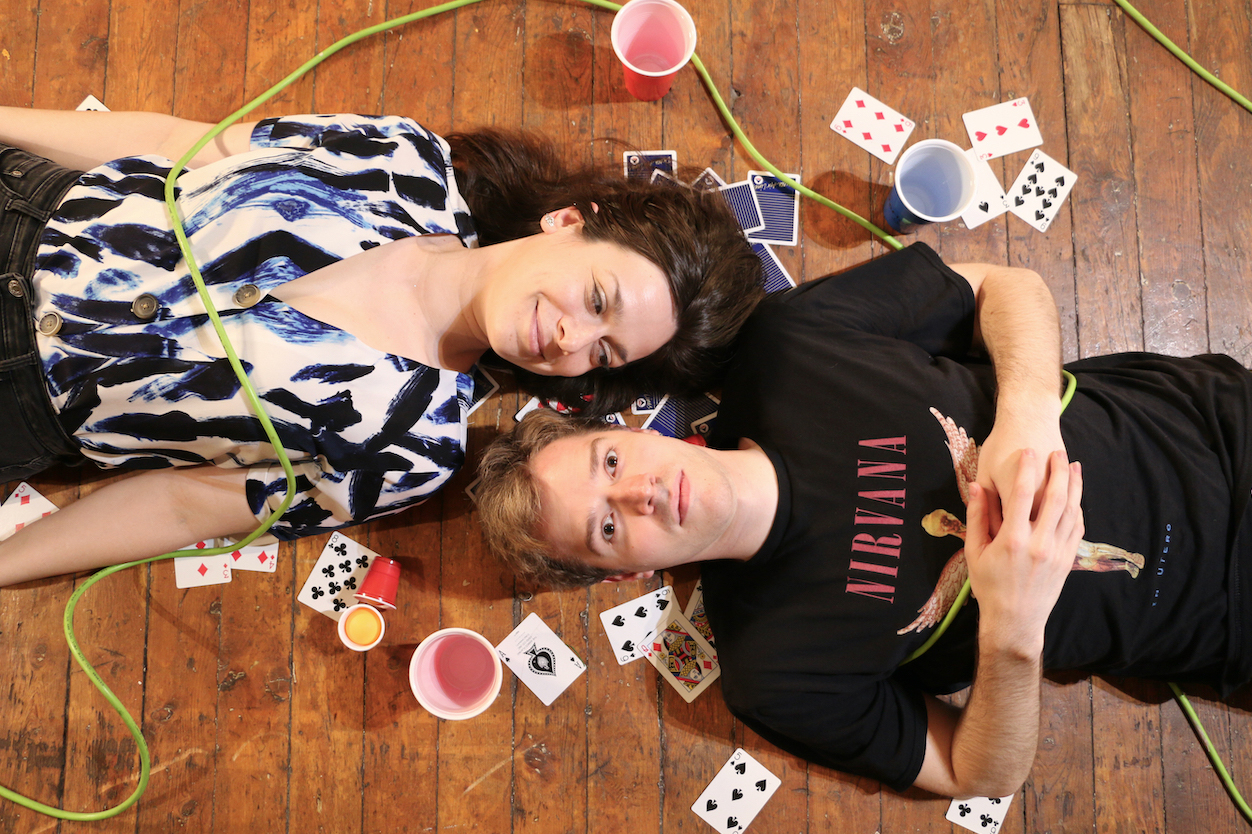 A couple lie on the ground of an old floor in a romantic but concerning pose. They are surrounded by junk, party cups, and electrical cords.