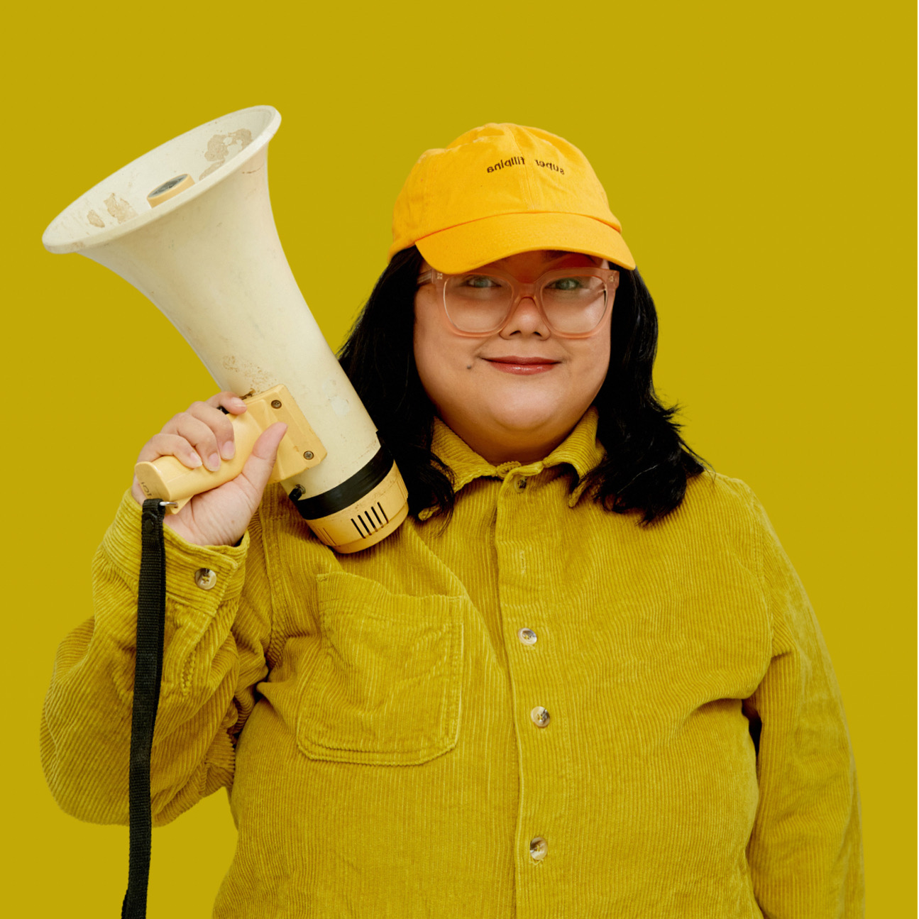 Alia is a Filipina woman standing in front of a mustard yellow background. She has black, shoulder-length hair, and is wearing glasses with light pink, transparent frames. She is wearing a mustard yellow corduroy buttoned up shirt. She is wearing a bright yellow cap that says “super filipina” in black. She is smiling and holding a megaphone.