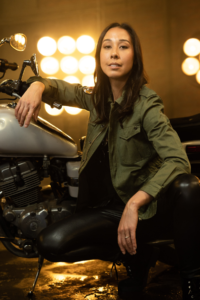 Flora Le, crouching on the side of a parked motorcycle, her back straight and right arm resting on the handlebars, looking at the camera.