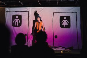  Grumms, a white non-binary performer with blue hair stands in front of a
screen in an orange bathing suit. On the screen, hand drawn images of a men’s and women’s
toilet doors are projected. They stand between the two doors, staring out at the audience. The
lighting is dim, pink and spooky. 